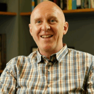 Matthew Taylor smiling at the camera in his office. Books are visible on the shelfs behind.