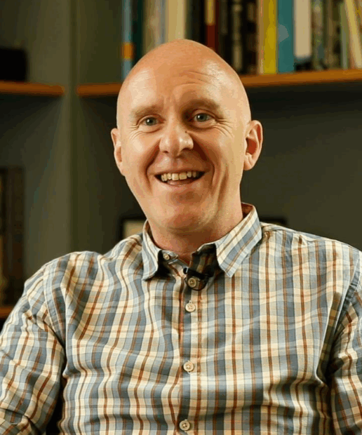 Matthew Taylor smiling at the camera in his office. Books are visible on the shelfs behind.