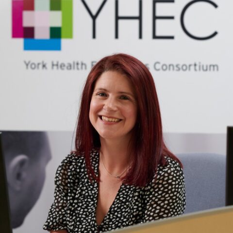Rachael McCool smiling at the camera in an office setting, with the YHEC logo visible on the wall behind her