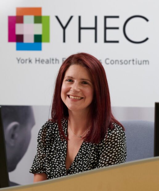 Rachael McCool smiling at the camera in an office setting, with the YHEC logo visible on the wall behind her