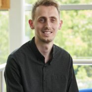 Rob Malcolm sitting on the edge of a sofa, smiling at the camera. Treese are vivible through the windows behind him