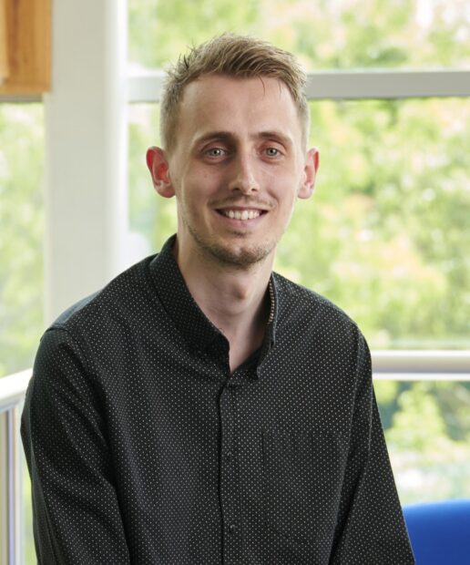 Rob Malcolm sitting on the edge of a sofa, smiling at the camera. Treese are vivible through the windows behind him