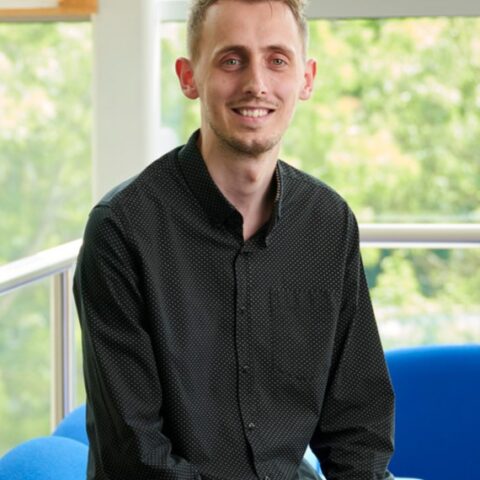 Rob Malcolm sitting on the edge of a sofa, smiling at the camera. Greenery is vivible through the windows behind him