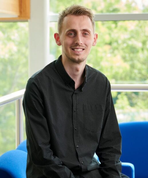 Rob Malcolm sitting on the edge of a sofa, smiling at the camera. Greenery is vivible through the windows behind him