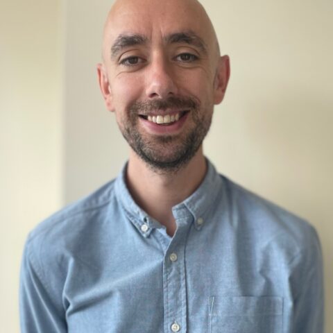 A head-and-shoulders photo of Will Green, smiling at the camera. He is stood against a neutral, cream background
