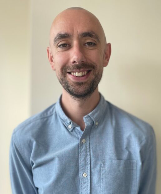A head-and-shoulders photo of Will Green, smiling at the camera. He is stood against a neutral, cream background