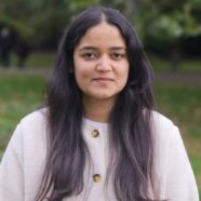 A head-and-shoulders photo of Angel. She is stood facing the camera and smiling. She is stood outside, with trees visible in the background