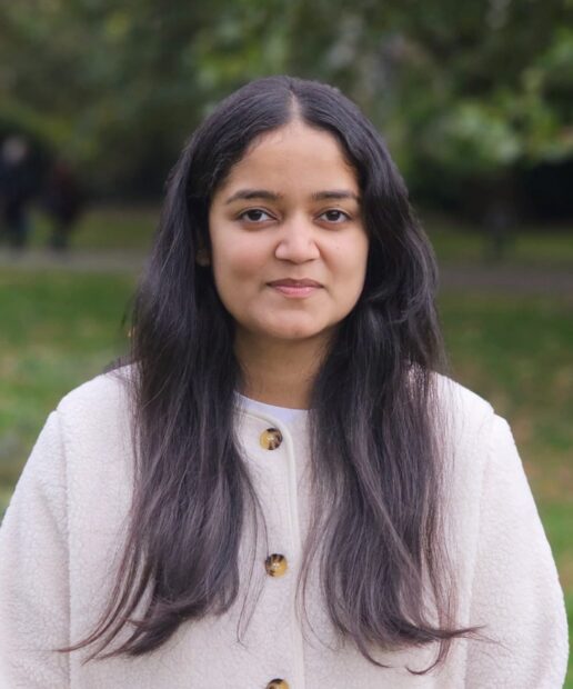 A head-and-shoulders photo of Angel. She is stood facing the camera and smiling. She is stood outside, with trees visible in the background
