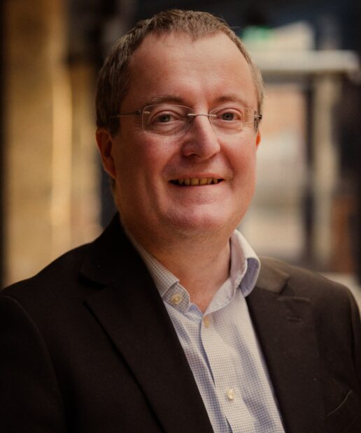 A portrait, head-and-shoulders image of Ken Badcock smiling at the camera. He is outside, with buildings visible (but blurred) behind him