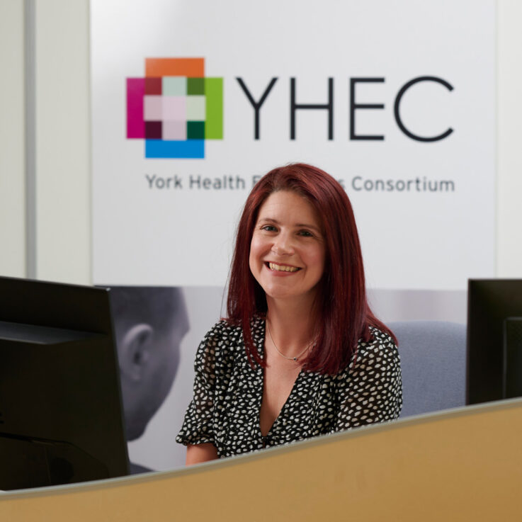 Rachael is sat behind a computer desk, smiling at the camera. The YHEC logo is visible on the wall behind her.