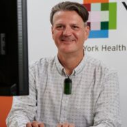 Hayden Holmes is smiling at the camera. He is sitting behind a desk, with the computer visible on the edge of the screen. The YHEC logo is visible on the wall behind