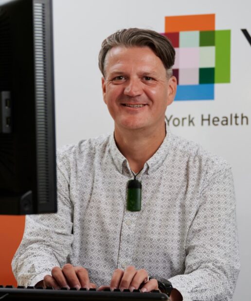Hayden Holmes is smiling at the camera. He is sitting behind a desk, with the computer visible on the edge of the screen. The YHEC logo is visible on the wall behind