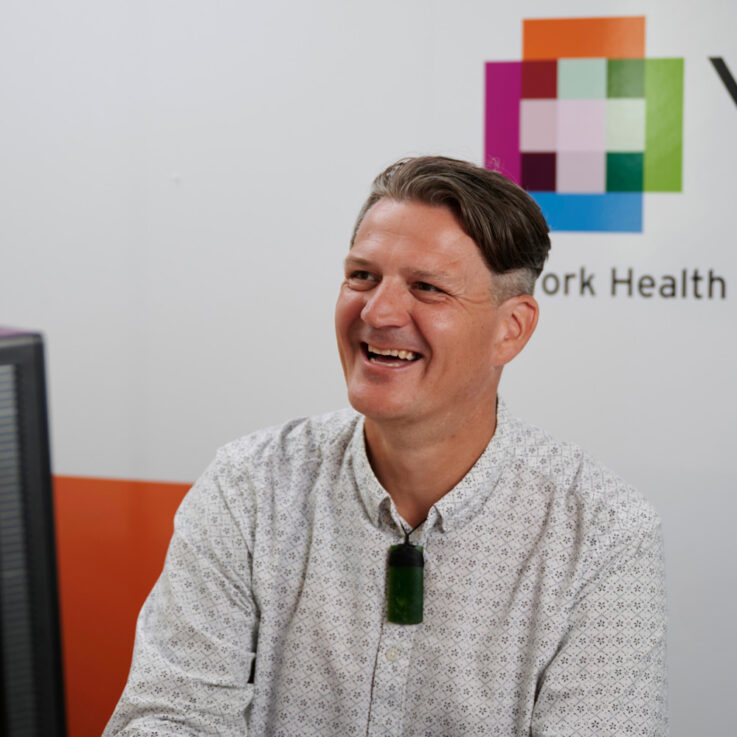 Hayden is sat behind a computer desk, looking and smiling at someone off-camera. The YHEC logo is visible on the wall behind him.