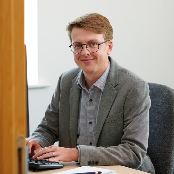 Joe Moss is sat behind a desk with his hands on the keyboard, smiling at the camera. The photo is taken through the door frame of the office, at an angle to the desk