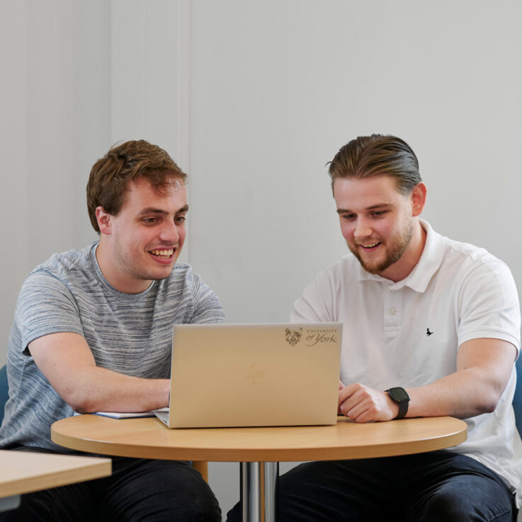 Two male workers sitting in an office, smiling and looking at a laptop. The photo is taken through the doorway of the meeting room
