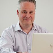 Nick Hex smiling, looking just off camera. He is sat in an office behind a desk, with a laptop in front of him