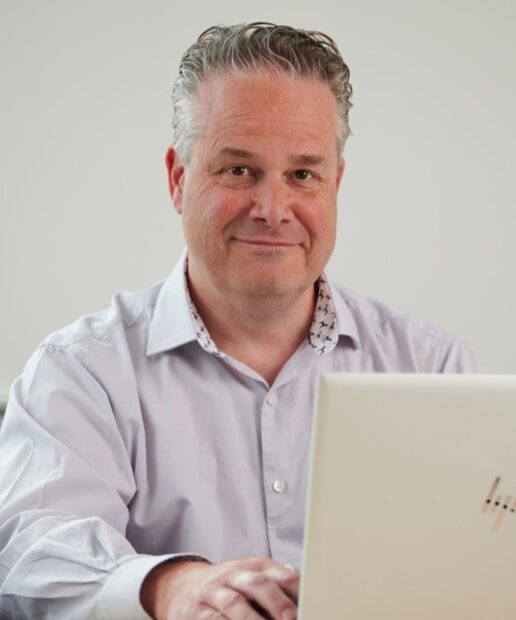 Nick Hex smiling, looking just off camera. He is sat in an office behind a desk, with a laptop in front of him