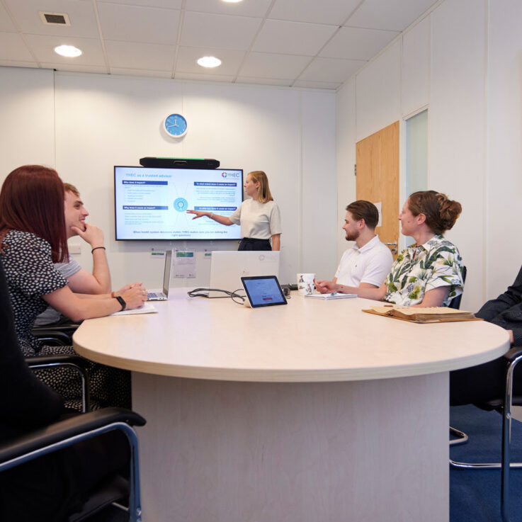 A woman presenting to a meeting room of 6 other people. The photo is taken from the back of the meeting room, and the presenter is gesturing towards the screen