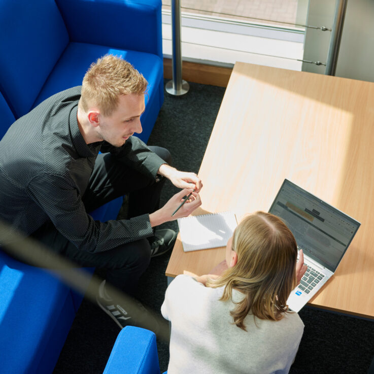 Two people sitting on blue sofas in an open space, talking with each other and leaning over a laptop and notebook