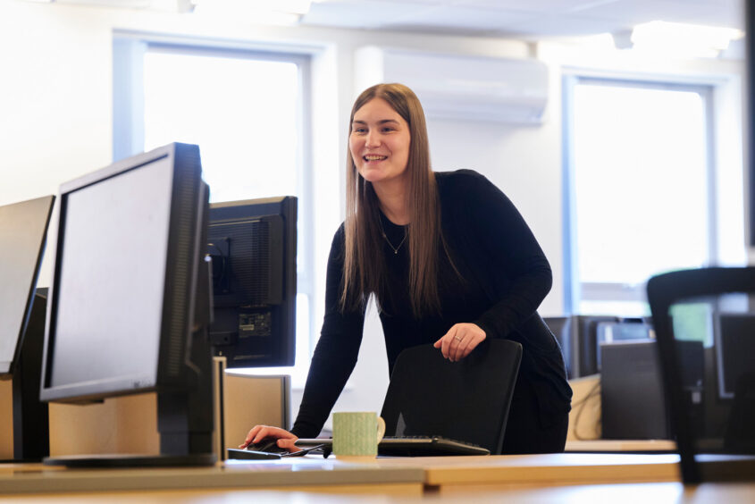 A woman in an office setting, standing behind a desk. She is leaning down to use the computer mouse, while talking to someone off camera