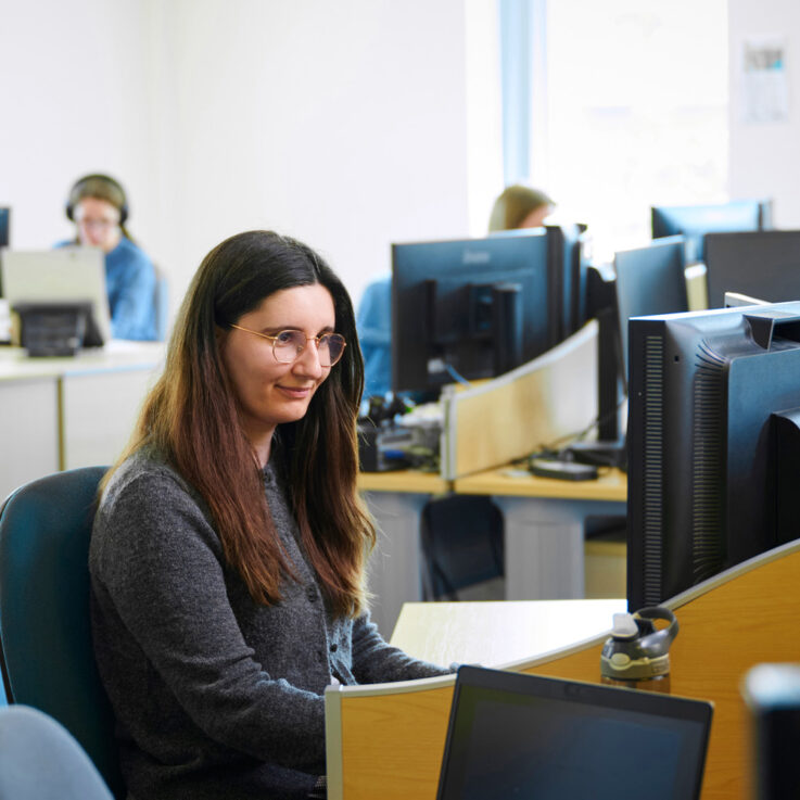 A female worker sitting at a desk, looking at the computer in front of her. Other computers and workers are visible in the background.