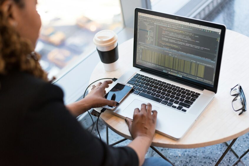 A laptop is facing the camera, at an angle. The laptop is sitting on a small table and is next to a coffee cub on one side and a pari of glasses on the other. A woman is sitting down at the edge of the photo, leaning over the laptop and working.