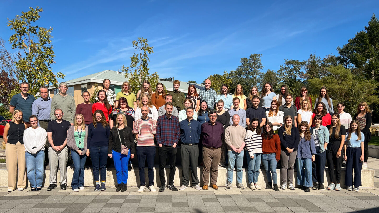 YHEC's annual, 2025 all-staff photo. The group of -60 indivudals are stood outside (on a sunny day) in a tiered format split over 3 rows