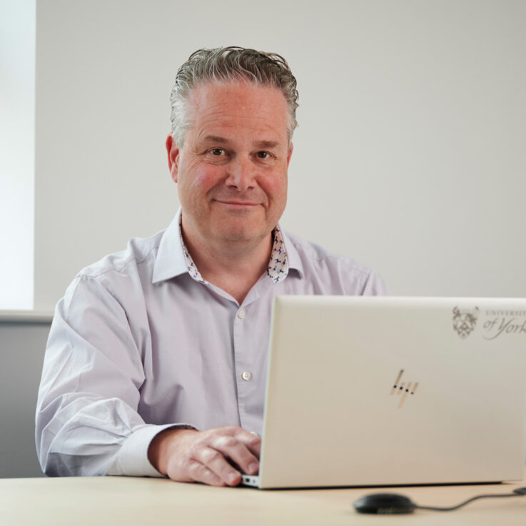 Nick Hex smiling, looking just off camera. He is sat in an office behind a desk, with a laptop in front of him